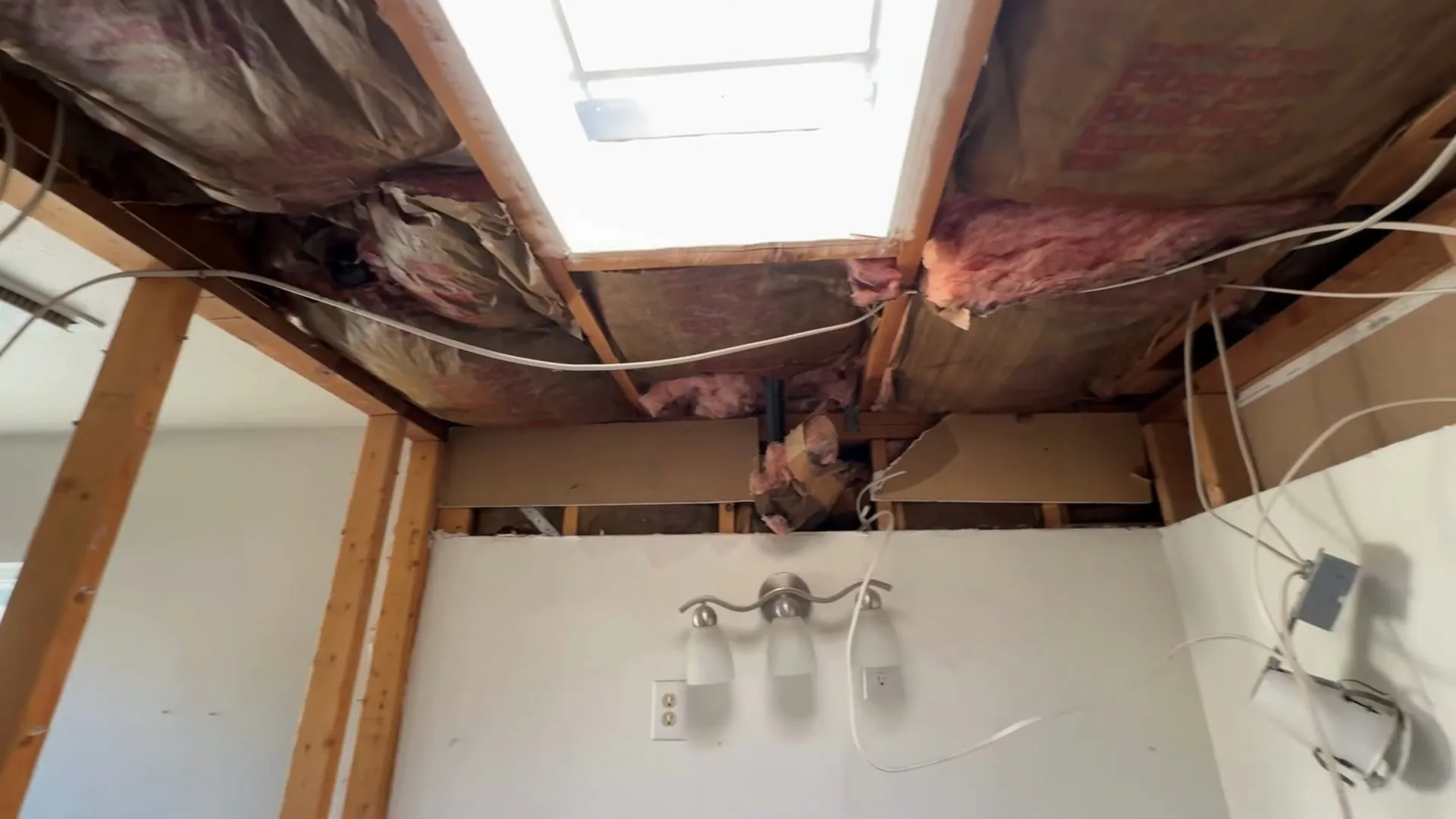Ceiling opened up to the joists, with pink insulation pulled down around a bright skylight opening and a cable hanging below