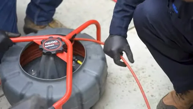 Technician crouching beside a grey camera reel with a red push rod, gloved hands on the cable