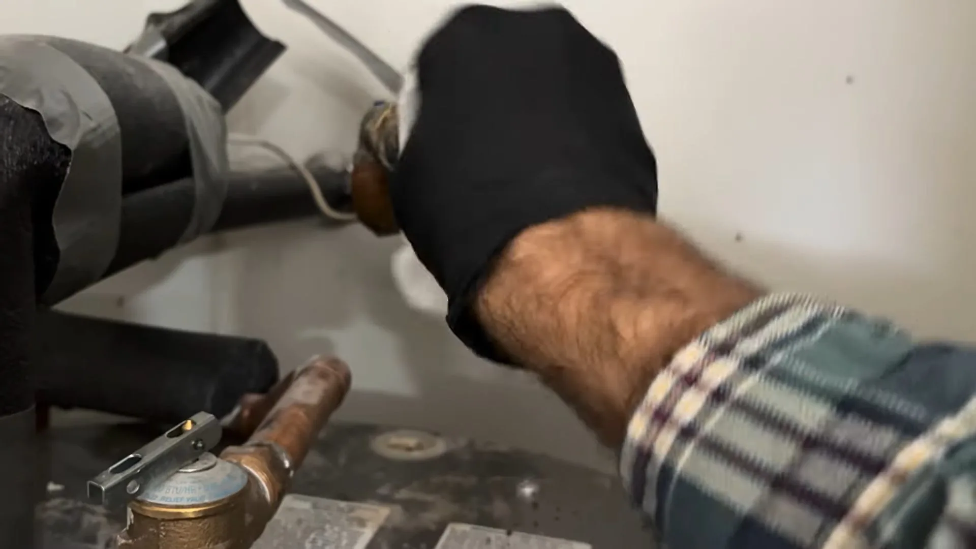 Hand in a black glove reaching to a copper fitting on top of a water heater beside lagged pipe