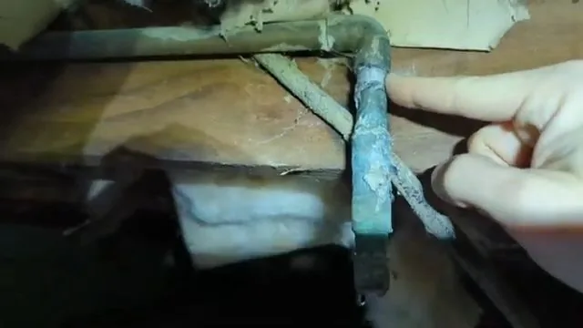 Hand pointing at a green corroded bend in a copper supply pipe under floor joists in a crawlspace