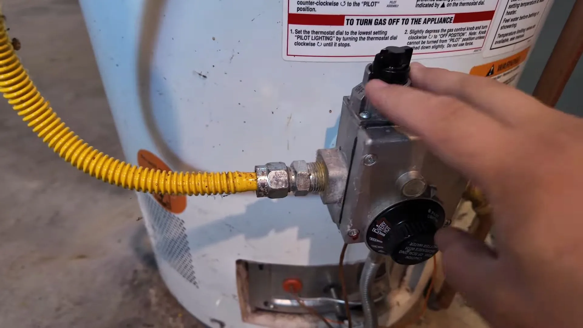 Hand on the gas control and pilot knob of a water heater with a yellow flexible gas line attached