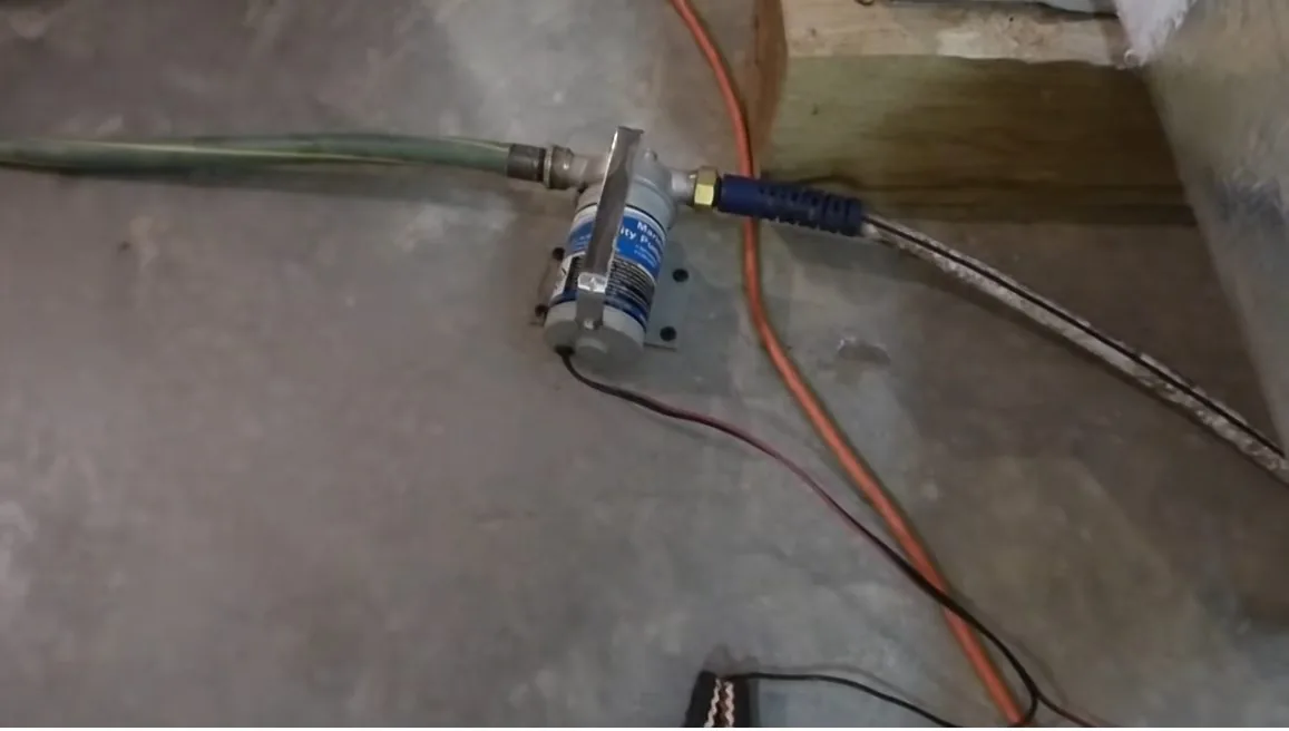 Small metal pump with a green hose attached, standing on a bare grey basement floor with an orange cord running away