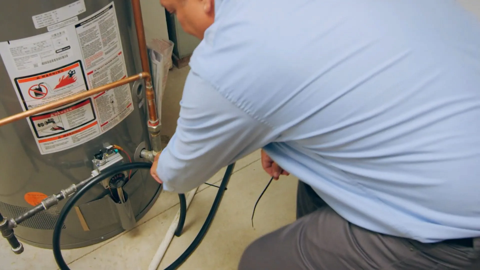 Technician crouched beside a gas water heater connecting a hose to its fittings