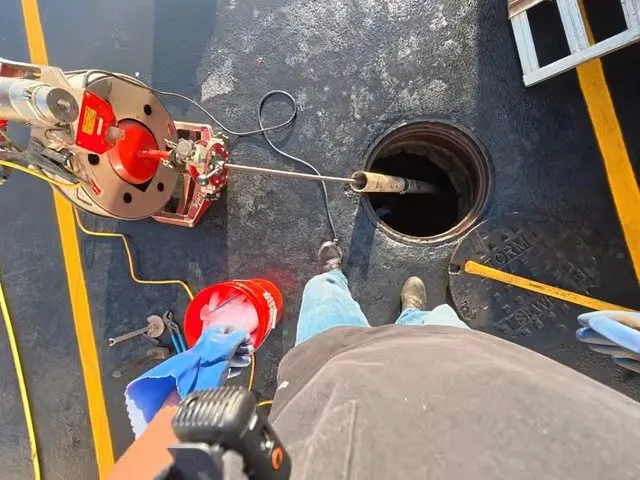 Overhead view of a technician feeding a red cable into a drain pit with a camera case beside him