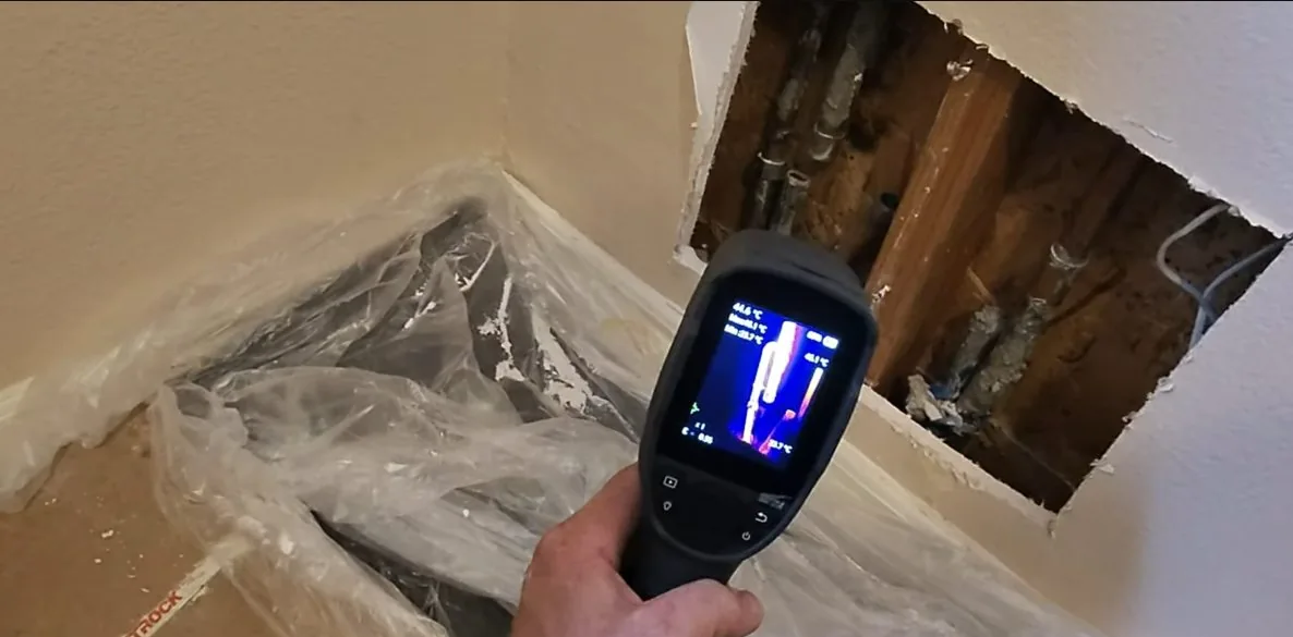 Hand holding a thermal imaging camera whose screen shows a warm vertical pipe, beside an opened wall cavity with copper lines
