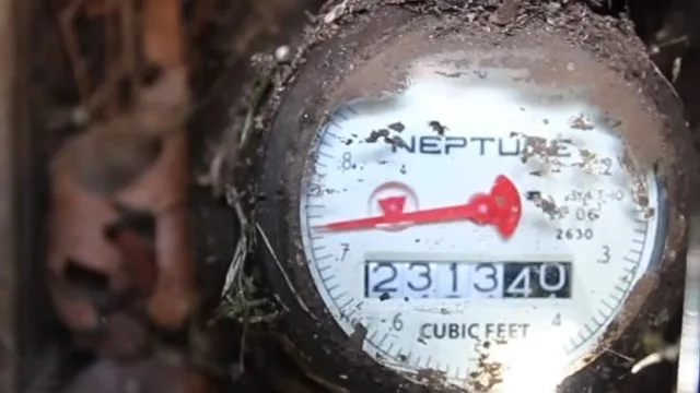 Water meter dial with a red sweep hand and a cubic feet register, set in a dirt-lined meter box