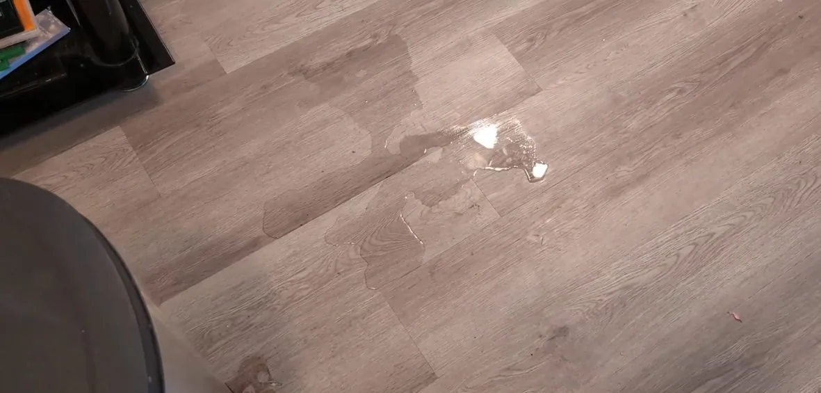 Shallow pool of water and darker damp patches spreading across a grey wood-effect floor